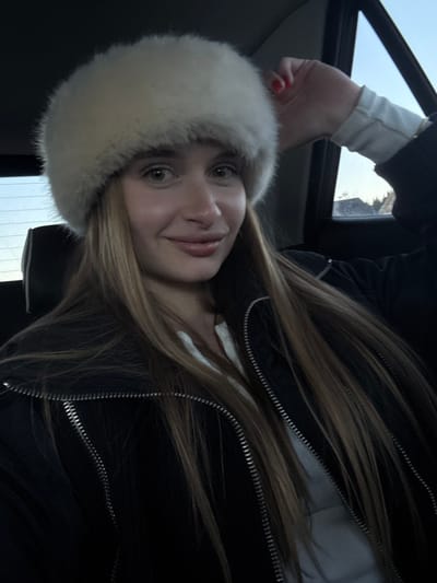 A female individual is seated in the rear passenger compartment of a vehicle. She wears a large, light-colored fur hat, a black jacket with silver zippers, and a white ribbed long-sleeved top. Her long, straight, light brown hair extends below her shoulders. She looks directly forward, exhibiting a slight smile. Her right hand, displaying red nail polish, is raised with fingers slightly curled, positioned near the vehicle's window frame. The headrest of the seat behind her is visible. Through the vehicle's side window, an outdoor scene is visible, including a light blue sky and dark shapes resembling distant trees or mountainous terrain, consistent with a winter or cold-weather environment. The image is described as being taken in Razvršje, Montenegro.