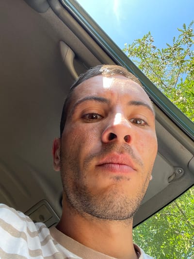 A man is captured in a selfie from a low angle, looking upwards. He is inside a vehicle, with the interior of the car visible in the foreground. Sunlight streams in, illuminating his face and the car's ceiling. Through the open window or sunroof, lush green trees and a clear blue sky are visible, suggesting an outdoor setting. The man is wearing a striped t-shirt. The overall scene appears to be a candid moment, possibly during a drive on a sunny day in the city of Levski, Bulgaria.
