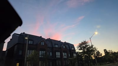 The media file depicts a street view of a residential building with a streetlamp and trees against a twilight sky. The building is a multi-story brick structure with dark-colored sloped roofs and multiple windows. A streetlamp with its light on stands to the right of the building, illuminating the surrounding area. Trees are visible in front of and to the right of the building, with some silhouetted against the sky. The sky is a gradient of blue and pink, indicating that it is either sunrise or sunset. The overall mood is peaceful and serene. The location is Vaughan, Canada. No visible text is present in the media file.
