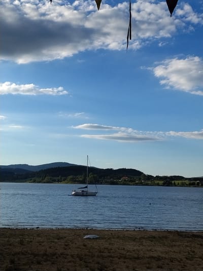 This is a vertical, eye-level, outdoor photograph captured on a sunny day, likely during the late afternoon given the lighting and cloud formations. The image depicts a tranquil scene at the Slapy Reservoir in Radslav, Czechia.
In the foreground, a dry, pebbly shore leads down to the calm, blue water of the reservoir. The water is rippled, suggesting a light breeze. There is a small, white object on the shore, possibly a piece of debris or a covered item.
The middle ground is dominated by the expansive reservoir. A white sailboat with a blue canopy is anchored or moored in the water, its mast reaching towards the sky. The boat appears to be unoccupied. Further in the distance, on the far side of the reservoir, is a dense, dark green treeline that forms a rolling hillside. Scattered amongst the trees are hints of buildings, suggesting a small settlement. The distant hills are bathed in a soft, muted light.
The sky above is a vibrant blue, with scattered white and gray clouds. Some of the clouds are cumulus, with soft, billowy shapes, while others are thinner and more wispy, stretched across the horizon. At the very top of the image, the dark shapes of triangular banners or flags are visible, dangling from an unseen structure above. These banners are a stark contrast to the bright sky and natural landscape.
The overall atmosphere of the image is one of peacefulness and serenity, with the natural beauty of the landscape and the solitary sailboat creating a sense of calm.