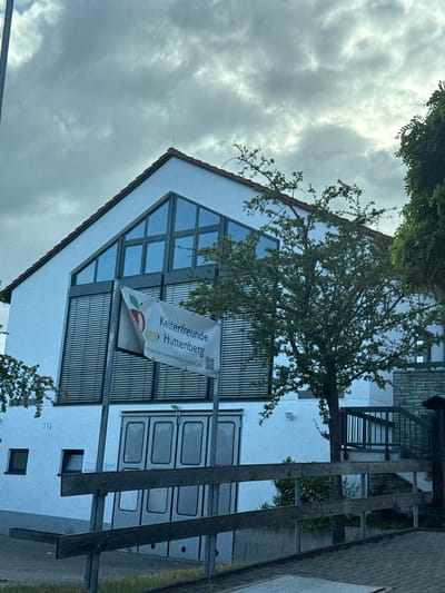 This media file shows the exterior of a white building with a triangular roof. A large tree with green leaves partially obscures the front of the building. A banner is hung on the building, featuring an apple graphic and text. The text on the banner reads "Kelterfreunde Hüttenberg" and "kelterfreunde-huettenberg.de". There are also QR codes visible on the banner. The building has several windows, some with dark blinds. To the right of the building, there is a fence or railing and some greenery. The sky is cloudy and overcast. A house number "114" is visible on the building. The scene appears to be a daytime shot in Volpertshausen, Germany. The overall mood is neutral, with no strong emotional cues conveyed.