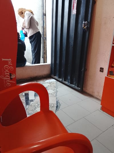 The image captures an indoor or semi-enclosed space, likely a waiting area or shop entrance, with a bright doorway leading to an outdoor or brightly lit area.
In the foreground on the left, a bright orange plastic chair is partially visible, showing its backrest and arm. To its left, a tall red vertical banner or board stands, displaying white text that reads "1st Eagle" prominently, and smaller text below that appears to say "O.T.G. Flash Memory Card".
In the midground, near the brightly lit doorway, two individuals are present. One person, a woman with light brown, curly, possibly braided hair, is standing with her back mostly to the viewer. She is wearing a light beige or off-white long-sleeved top and dark trousers. She is leaning forward, seemingly attending to something held within a blue plastic bag or container. Partially obscured by the red banner and the standing woman, another person is seen sitting or kneeling on a low step or the ground. This person is wearing dark clothing, and white text, "y Later!", is visible on their back, possibly part of a design on their shirt.
To the right of the doorway stands a dark-colored, vertically ribbed metal door or panel, which appears to be slightly ajar or positioned as a fixed element beside the main opening. Two small, shiny padlocks are secured to a latch on its right side. A small red sticker with white text, partially visible as "inder", is affixed to the upper part of this door.
The floor inside the room features light-colored square tiles. Closer to the doorway, there's a section of patterned tiles with geometric shapes in varying shades of grey, white, and possibly light brown. The wall on the right side of the image is painted a light peach or orange color, and a dark, square-shaped opening or mark is visible on it. Bright light streams in from the open doorway, illuminating the scene and suggesting it is daytime.