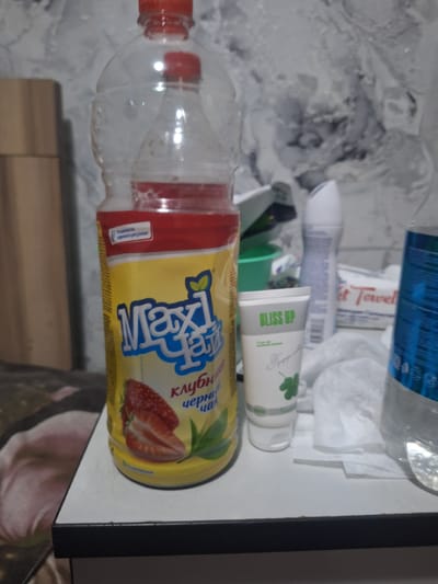 The image displays a close-up view of a white countertop with various items arranged on it. In the foreground, on the left, is a large, partially empty plastic bottle with a red cap, containing a yellow liquid. The label on the bottle is predominantly yellow and features the text "Maxi" and an image of strawberries, along with Cyrillic text. To the right of the bottle, there is a small white tube with green text that reads "BLISS UP." Behind these items, a white spray can with blue accents and a white, textured bottle are visible, along with a stack of white paper products. A blue-tinted plastic bottle is present on the far right. The background is a grey, textured wall with a mottled pattern. The lighting appears to be artificial, casting subtle shadows. The scene is static and shows no signs of activity. The location is identified as Ötegen batyr, Kazakhstan.