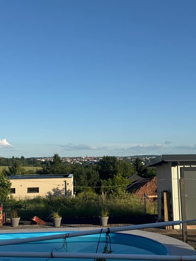 This image captures an outdoor residential scene with a panoramic view of the city of Pirmasens, Germany, under a clear, bright blue sky featuring a few scattered white cumulus clouds.
In the immediate foreground, an oval-shaped, above-ground swimming pool with a blue liner is partially covered by a light grey tarp. A white PVC pipe or bar spans horizontally across the pool's surface, with dark ropes or straps hanging from it. To the left of the pool, three grey cylindrical planters containing green plants are positioned on dark grey pavers. Between two of these planters, a red, triangular object rests on its side.
Behind the pool area, a metal fence separates the foreground from the midground. To the left, a light tan, modern building with a flat roof is visible, featuring multiple solar panels and two narrow, horizontal rectangular windows. Sunlight casts shadows on its facade. To the right, part of a light-colored, vertically corrugated utility shed or building is present, characterized by a dark roof overhang and a dark grey door. A wooden post is leaned against its exterior. Behind this building, a large pile of reddish-brown earth is evident.
The midground is dominated by a dense band of green trees and overgrown vegetation. Beyond this tree line, expanses of green fields are visible.
The background reveals the city of Pirmasens, Germany, stretching horizontally across a distant hill. The cityscape comprises numerous residential and commercial buildings, predominantly with dark roofs. A distinctive church steeple or tower with a reddish-brown spire is visible near the center-right of the urban profile. The city is framed by rolling, forested hills. No people or discernible actions are present in the scene.