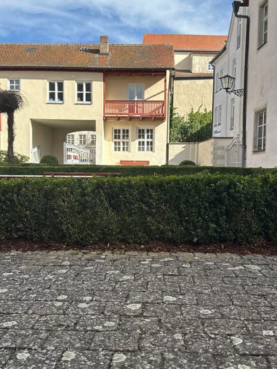 The image displays an exterior daytime scene in Weißenburg in Bayern, Germany, featuring historical European architecture and landscaping under a partly cloudy sky. The foreground consists of a grey paver pathway exhibiting lichen growth. Immediately behind this, a neatly trimmed dark green hedge with brown mulch at its base extends horizontally across the frame. A reddish-brown wooden park bench is partially visible behind the hedge.
In the background, a row of interconnected two-story buildings dominates the scene. The central structure features a light yellow stucco facade, a reddish-brown tile roof, and a prominent red-painted wooden balcony on the first floor with a white-framed window/door opening onto it. Below the balcony, two white-framed windows are present on the ground floor. To the left of this central section, the building includes an arched ground-floor passageway leading into a courtyard, where a white metal gate is visible. Two white-framed windows are on the first floor to the left of the archway. A bare, dark-branched tree stump is partially visible on the far left. A chimney stack protrudes from the roofline of the central building.
To the right, a taller, multi-story building with a light cream-colored facade stands adjacent, featuring an ornate black metal lantern affixed to its wall and multiple rectangular windows. A stone wall with a white metal gate continues from the courtyard entrance area. Green foliage grows against a lighter wall visible between the central and right-most buildings. Strong sunlight illuminates the scene, casting distinct shadows from the buildings and architectural features. No people or discernible actions are present.
