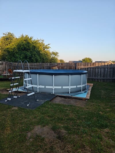 The image shows an above-ground swimming pool set up in a backyard. The pool is round with a grey exterior and a blue cover. A silver ladder with white steps is positioned to the left of the pool. The pool is situated on a wooden platform, which is placed on a dark mat. The backyard is grassy, with a wooden fence surrounding it. In the background, there are trees and some buildings. The sky is clear and blue. The overall scene suggests a residential outdoor area, likely a private property in the village of Garochytsky Sel'ski Savet, Belarus.