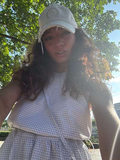 A medium, angled shot depicts a person with brown curly hair and a white cap, wearing a white and black checkered top, likely in an outdoor setting. Green foliage from trees is visible in the background along with bright sunlight and patches of blue sky. The location is Lao Cai Ward, Vietnam.