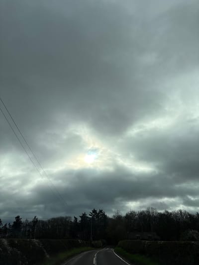The image shows an overcast sky with the sun faintly visible through the clouds. Below the sky is a road lined with trees and vegetation on the right side. Two utility wires stretch diagonally across the lower left of the frame. The scene appears to be located in a rural or semi-rural area, possibly near Baschurch, United Kingdom, based on context.