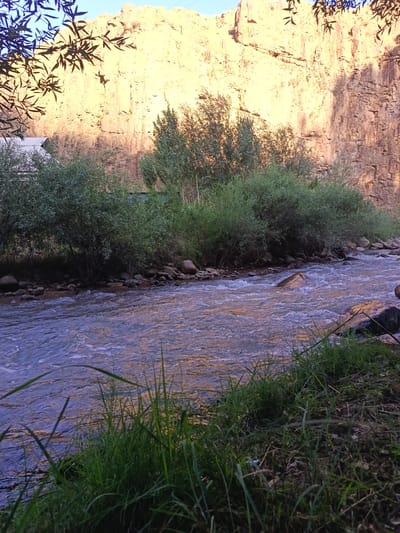 The image depicts a natural scene featuring a flowing river in the foreground, bordered by lush green grass and vegetation on the right bank. The river water is a deep blue-purple, with whitecaps indicating movement. The left bank of the river is lined with trees and bushes, behind which a small white structure, possibly a building or shed, is partially visible. In the background, a large, rocky cliff face dominates the scene, illuminated by warm sunlight, casting shadows. The cliff is characterized by its rough, textured surface and geological striations. Tree branches with dark leaves are silhouetted against the bright sky in the upper left corner. The overall environment suggests a rural or semi-natural setting, potentially on the outskirts of a populated area. The location is identified as Akhmatabad, Uzbekistan. There are no people or animals visible in the image. The scene appears to be captured during daylight hours, with the sunlight suggesting either early morning or late afternoon.