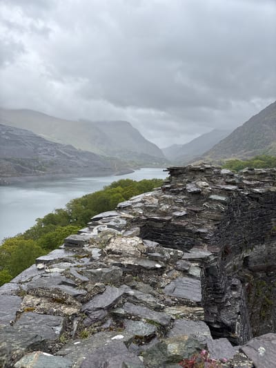 The image presents a scene in Llanberis, United Kingdom, likely a landscape view from a high vantage point. In the foreground is a rugged rock face composed of dark, layered stone. Green vegetation, possibly trees or shrubs, lines the bottom of the rock face. In the mid-ground, a body of water stretches into the distance, with a rocky mountain rising on the left side of the water. The sky is overcast, with dense grey clouds obscuring the upper portion of the mountain, creating a misty atmosphere. The image is rotated counterclockwise.