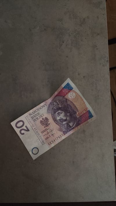 A Polish 20 zloty banknote is lying on a grey, textured surface, possibly a countertop or table. The banknote is angled, with the left side higher than the right. The denomination "20" is visible in large purple numerals on the left. The text "NARODOWY BANK POLSKI" is printed in Polish on the banknote, along with the image of a historical figure, likely Bolesław Chrobry, the first king of Poland. A serial number, "CE5417621", is also visible. The banknote appears to be in good condition. The background is a dark, neutral grey with some subtle variations in tone and texture. The environment does not provide any specific clues to the city of Sosnowiec, Poland.