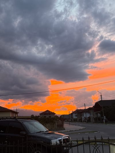 The image captures a dramatic sunset over the town of Blatska, Bulgaria. The sky is a brilliant spectacle of orange and red hues, with dark, heavy clouds dominating the upper portion of the frame. In the foreground, a black SUV is parked behind a black metal fence. The vehicle appears to be a mid-size SUV, possibly modified with a roof rack. The street is paved, with sidewalks and houses lining one side. The architecture suggests a European setting. A lamppost is visible on the right side of the image. The overall scene conveys a sense of a peaceful evening in a residential area, with the natural beauty of the sunset providing a striking backdrop.