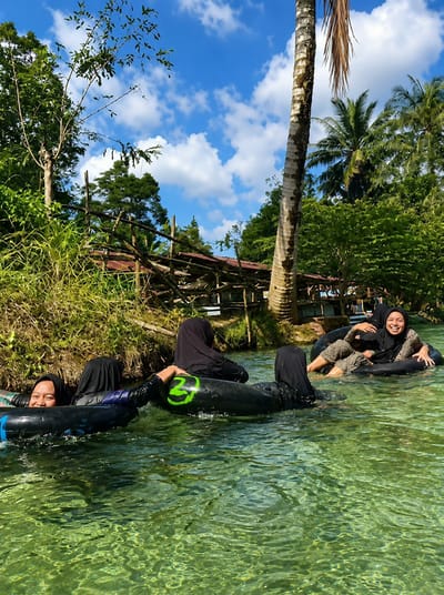 Four individuals, three women and one person whose gender is not discernible, are depicted floating in clear, greenish water on inner tubes. They are all wearing dark-colored clothing, with three individuals wearing hijabs. The woman in the foreground on the left is looking towards the camera and smiling. The woman in the background on the right is also smiling and looking towards the camera, with her legs crossed. The water is shallow enough to see the sandy bottom. The background features lush green vegetation, including trees and tall grasses, with a wooden structure resembling a fence or a simple building visible behind them. A tall palm tree stands prominently on the right side of the frame. The sky is blue with scattered white clouds. The scene appears to be a recreational activity in a natural outdoor setting, possibly a river or a clear spring in Silaiyajae, Indonesia.