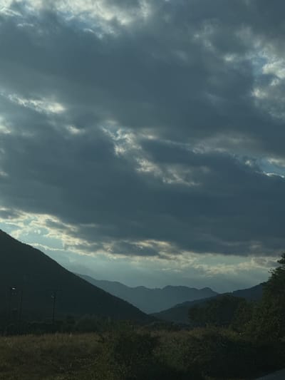 The image captures a dramatic, moody sky over a mountainous landscape. Heavy, dark clouds dominate the upper portion of the frame, with shafts of light breaking through in the distance, illuminating the layers of mountain ridges. The foreground features the dark silhouette of a sloping hillside covered in dry grass and vegetation. Two utility poles are visible on the hillside. In the far distance, more layers of mountains fade into a hazy blue. The overall impression is one of a pre-storm atmosphere. The location is identified as Melivoia, Greece.