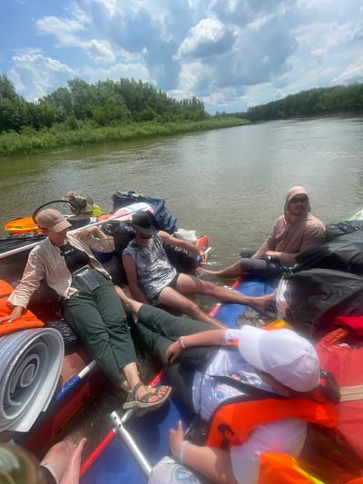 Four individuals are seated on a raft on a wide, calm river. The raft is laden with various bags and equipment, including orange life vests and rolled-up mats. The surroundings are lush and green, with dense trees lining the riverbanks and a partly cloudy sky overhead. The scene appears to be a recreational outing, possibly a river trip, in or around the settlement of Nezhinka, Russia, given the vegetation and river type common in the region.