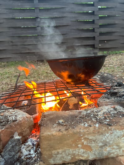 The image depicts a fire pit constructed of bricks and containing burning wood. A metal grill sits atop the fire, and a dark, rounded metal pot is positioned on the grill, emanating smoke towards a grey wooden fence in the background. The scene is likely outdoors, with grass visible in the background. The image does not include specific indicators of being in Новоклязьминское, Russia. There are no people visible in the image.