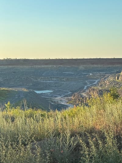 The image depicts an aerial view of a quarry with tiered walls, likely a mining operation, in Tatsinskaya, Russia. The quarry floor is uneven, with some areas showing water accumulation, forming a small, shallow pool. Lush green vegetation lines the upper edges of the quarry, cascading down in some areas. The sky is a soft gradient of light blue transitioning to pale yellow, suggesting it is either early morning or late afternoon. The lighting casts a warm glow on the vegetation, highlighting its texture. There are no people or explicit man-made objects visible other than the quarry itself. The overall scene conveys a sense of scale and the raw, natural beauty of an industrial landscape interacting with the environment.