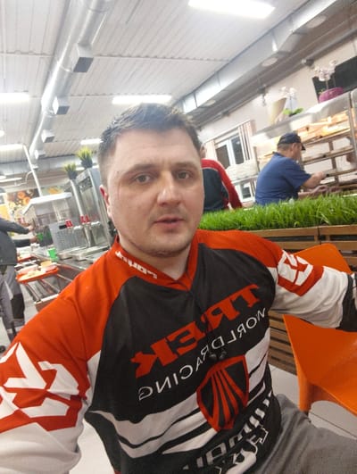 The image depicts an indoor scene in the city of Im. Gastello, Russia, appearing to be a cafeteria or self-service restaurant. The environment features a corrugated white metal ceiling with exposed silver ventilation ducts and bright overhead fluorescent lighting.

In the foreground, a light-skinned adult male with short, dark hair and a neutral expression is visible, facing directly towards the camera, suggesting a selfie. He wears a long-sleeved athletic jersey, predominantly black and orange with white geometric patterns and text. The front of the jersey displays "TREK WORLD RACING" in white capital letters, with a red stylized emblem below. He is also wearing grey pants, and a segment of an orange plastic chair is visible on his right.

The mid-ground features a decorative planter containing green, grass-like artificial plants, acting as a visual divider. Behind this planter, to the right, an adult male wearing a dark blue short-sleeved shirt and a dark baseball cap is actively working behind a food service counter, appearing to be arranging or preparing food within a heated display unit. Further to his left and in the background, another individual in a red and black jacket is partially visible near stacks of white bowls. To the far left background, the lower half of an individual's legs, clad in grey and black pants, is visible near a counter with metal trays holding food items and multiple beverage dispensers. Various decorative elements, including potted succulents and flowers, are present on shelves in the background.