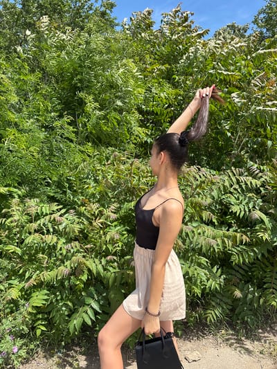 A young woman stands outdoors near dense green foliage. She wears a black top, a light-colored skirt, and has her dark hair pulled back in a ponytail. She raises her right arm, holding the end of her ponytail in her hand. Her left hand holds a black purse. The location is presumed to be Mihailovo, Bulgaria, based on the prompt's instructions. The immediate environment is characterized by lush greenery.