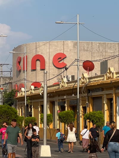 The image shows a street scene in San Salvador, El Salvador. The "CUC" signage on a building facade suggests the location might be near a Centro Universitario Católico. The building architecture features classical detailing with decorative moldings. A red circular structure, possibly a loudspeaker or some type of signal device, is mounted on the building. Below, a Subway restaurant signage is visible. A variety of people are walking on the sidewalk, including a woman in a white dress, a group of younger individuals, and several other adults in casual clothing. Trees and bushes line the sidewalk. Overhead, utility lines and a lamp post crisscross the sky.