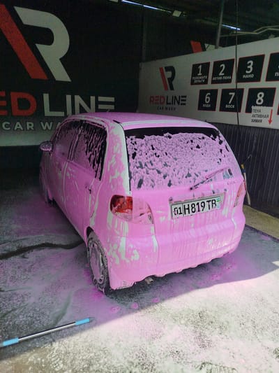 This media file captures a pink car covered in pink foam at a car wash. The car, a hatchback, is positioned on a wet, foamy surface, suggesting it's in the midst of a wash cycle. In the background, a prominent sign for "REDLINE CAR WASH" is visible, with numbered service options in Russian, such as "ПОЛОЖИТЕ ДЕНЬГИ" (Deposit Money), "АКТИВ ПЕНА" (Active Foam), "НАНО" (Nano), "ВОДА" (Water), "ВОСК" (Wax), and "ПЕНА АКТИВНАЯ ХИМИЯ" (Active Foam Chemistry). The car has a license plate that reads "01 H819 TB UZ", indicating it is from Uzbekistan. The lighting suggests it could be daytime, possibly overcast or indoors at a car wash. The scene appears functional and clean, despite the foamy mess. The overall impression is a typical car wash scenario, with a bright, eye-catching pink foam used for the cleaning process.