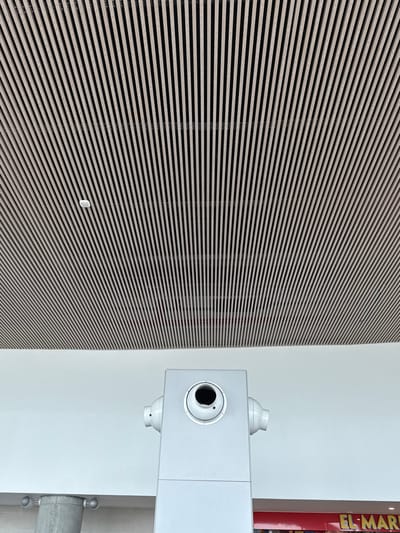 The scene is located in the city of AMCO, Area Metropolitana Centro Occidente, Colombia.
The image presents an indoor architectural perspective, dominated by a diagonally slatted ceiling composed of light-toned rectangular elements. A small, white, circular sensor or detector is affixed to this ceiling in the upper-left quadrant. Below the ceiling, a clean white wall occupies the middle section of the frame. A prominent light-gray, rectangular architectural element or column stands in the lower-center, extending upwards from the bottom edge. This element features a large circular recess with a dark, possibly empty, interior, and a smaller circular component offset to its right, suggestive of a camera or sensor housing. Two symmetrical, light-gray cylindrical fixtures protrude horizontally from either side of this rectangular element. To the left of this central element, a cylindrical concrete column is partially visible, featuring two circular lights or fixtures. In the lower-right corner, a section of a red sign with white capital letters, partially reading "EL MAR," is visible, indicating a commercial establishment. No individuals are present in the frame.