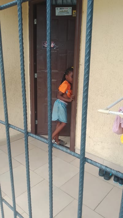 The image shows a young Black girl standing at a doorway, peeking around the edge of the doorframe into what appears to be a room. She is wearing an orange t-shirt that is pulled up, revealing her midriff, and a light blue denim skirt. She has her hair in braids with yellow ties. She is barefoot, wearing sandals. The doorway is dark brown and has some stickers on it. One sticker reads "2023 Unlimited POSSIBILITIES." Another sticker has "Ste ful" written on it. The girl's stance and the way she is peering suggest curiosity or a playful moment. The setting appears to be outdoors, possibly a porch or patio, with a tiled floor and a textured wall. In the background, a clothesline with some pink and yellow fabric is visible. Several pairs of black sandals are lined up on the floor near the wall. The lighting suggests it is daytime.