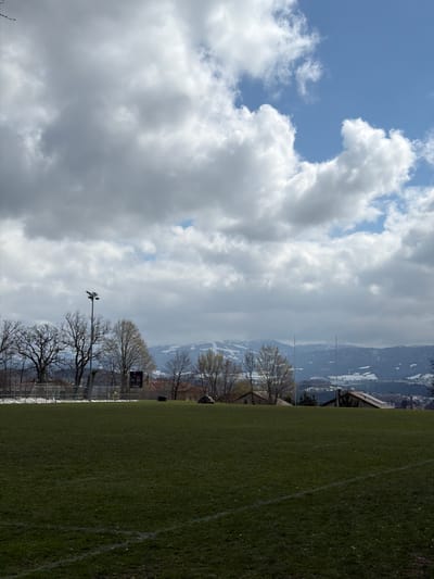 The image depicts an outdoor sports field, likely a football pitch, located in Fribourg - Freiburg, Switzerland, with snow-capped mountains in the distance under a dynamic, partly cloudy sky. The foreground features a dark green grass pitch with visible, faded white boundary lines.

In the midground, from left to right, a chain-link fence or netting is visible behind a goal structure. Adjacent to this, several leafless deciduous trees stand alongside others exhibiting nascent spring foliage. A tall, dark grey floodlight pole, equipped with multiple lamps, rises prominently. To its right, a black digital scoreboard is mounted on a pole. The scoreboard displays "24" on the left, "0:00" in the center, and "0" on the right. A red UBS logo is present on the upper left of the scoreboard, and the word "HUBLOT" is legible below the digital display. Further to the right, several residential buildings with reddish-brown tiled roofs are visible; one structure features rooftop solar panels. A large, dark, irregularly shaped object lies on the ground near these buildings. Two tall, slender white posts, resembling rugby goalposts, extend vertically against the distant landscape.

The background is dominated by a range of snow-dusted mountains, showing exposed rock and dark tree patches, indicative of a late winter or early spring season. The upper portion of the image displays a sky with large grey and white cumulus clouds partially revealing patches of clear blue sky.