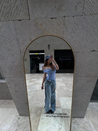 A full-length mirror in an arched gold frame stands outside a building in Ciudad López Mateos, Mexico. The mirror reflects a young woman standing and taking a selfie with a phone, partially obscuring her face. She is wearing a light blue top, jeans, and a blue hat. The reflection also shows the entrance to a business called "OM Clinic Center," and a currency exchange with reversed signage. The surrounding architecture consists of textured stone walls and tiled flooring. In the lower right corner of the mirror, the words "@omcliniccenter #tumejorversión" are displayed.