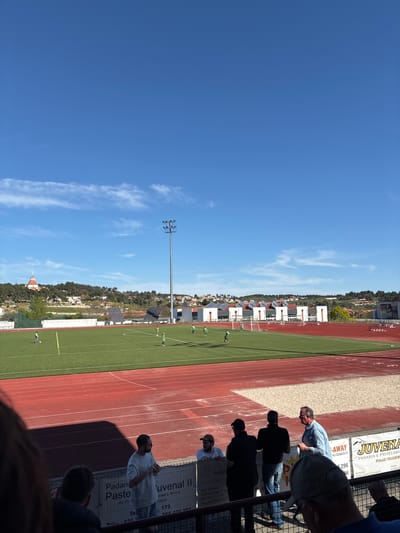 The image shows a soccer field and a track on a sunny day, viewed from the stands. A group of people watch the scene from behind a fence that is adorned with advertisement banners. The ads mention "Juvenal II," "Padari Paste," and a local "Sociedade," suggesting the field is in the municipality of Valpaços in Portugal, specifically in Valpaços, as indicated by "Sede: Valpaços" on one of the ads. Other elements in the background include houses, trees, and a lamp post, indicating an urban periphery. The sky is mostly clear with a few clouds.