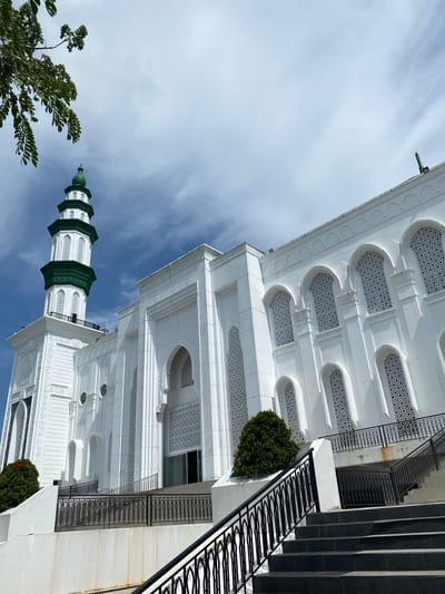 The image shows a white mosque with a green-domed minaret in Aluekambuk, Indonesia. The mosque features intricate geometric patterns on its windows and facade. A wide set of stairs leads up to the mosque's entrance, with black metal railings on either side. Lush green trees and foliage are visible around the base of the building. The sky above is blue with scattered clouds. There are no people visible in the image.