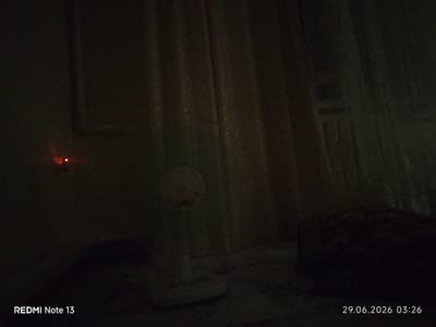 The image is a low-light, indoor scene. A white electric fan with a yellow center is positioned in the foreground, slightly to the left of center. Behind the fan, vertical curtains are visible, appearing to be made of a light-colored, sheer fabric. To the left of the fan, a small red light is illuminated on a dark surface, possibly an electrical outlet or indicator light. In the lower right quadrant of the image, a dark, textured object, possibly a piece of furniture or bedding, is visible. The bottom edge of the image displays a timestamp "29.06.2026 03:26" and a device identifier "REDMI Note 13". The environment is dimly lit, suggesting it is nighttime or the lights are off. No people or specific actions are depicted. The setting is indoors, likely a room in a residence.