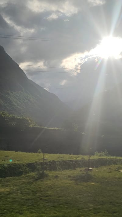 This is a vertical, medium-shot photograph of a mountain valley. The sun is bright and in the upper right corner of the frame, casting strong rays that create a lens flare effect across the image. Dark clouds are present in the sky, partially obscuring the sun. A steep, green, vegetated mountain slope dominates the left side of the image, disappearing into the misty valley. In the foreground, there is a grassy field with a gently sloping incline. There are power lines visible across the upper portion of the frame. No people or animals are visible. The location is specified as the city of Buron, Russia.