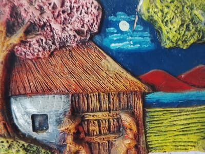 The image displays a detailed view of a three-dimensional artwork, specifically a painted relief sculpture or plaque, presenting a stylized nocturnal rural scene. This scene is identified as being located in Bernal Este, Argentina. The artwork is characterized by significant surface texture across all depicted elements.
In the mid-ground, a structure resembling a house or hut is positioned, featuring a textured, reddish-brown thatched roof with pronounced horizontal striations. The visible portion of the left wall is grey, containing a dark, square-shaped opening. Adjacent to this, a vertical, textured brown section represents a wooden door or wall panel. Above and to the left of the structure, a large, irregularly shaped, reddish-brown mass with a lumpy texture occupies the upper left quadrant, depicting foliage. A vertical yellowish-brown element, consistent with a tree trunk, is partially visible next to the house.
The background on the right side of the composition features a dark blue sky with a circular white moon and two distinct white cloud formations. Below the sky, two prominent, undulating red forms are present, depicting hills or mountains. A horizontal band of bright blue is situated beneath these red forms, representing water. The lower right foreground consists of a textured bright yellow field, exhibiting horizontal patterns. A small, light-colored, humanoid figure is partially visible at the bottom center-right, positioned between the house structure and the yellow field.