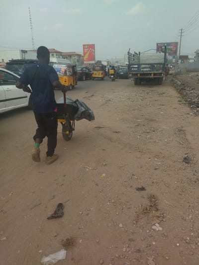 The image shows a man pushing a wheelbarrow on a dirt road in the city of Way, Nigeria. The coordinates provided locate the scene in the city of Way, Nigeria. The man is wearing a blue shirt, black pants, and brown shoes. He is pushing the wheelbarrow with his right hand and holding onto the handle with his left hand. The wheelbarrow is filled with a black tarp. The road is dusty and bumpy, with a few cars parked on the side of the road. There are several people walking on the sidewalk, including a woman in a blue dress walking towards the right edge of the image. The scene is set against a backdrop of buildings and a billboard. The sky is cloudy and hazy.