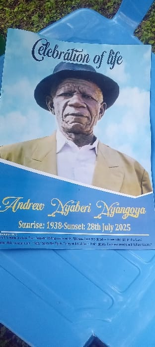 The image displays a "Celebration of Life" funeral program, likely resting on a light blue plastic chair or surface, with green grass visible in the background, suggesting an outdoor setting in Esise ward, Kenya. The program features a portrait of an elderly African male, identified as Andrew Nyaberi Nyangoya, wearing a dark fedora-style hat, a light-colored suit jacket, and a white shirt. The text indicates his birth year as "Sunrise: 1938" and his death date as "Sunset: 28th July 2025," followed by a partial quote from Revelation 14:13. The program is the primary subject, laid flat, indicating a pre-event setup or memorial context. No human interaction or specific action is depicted beyond the static arrangement of the program on the outdoor surface.