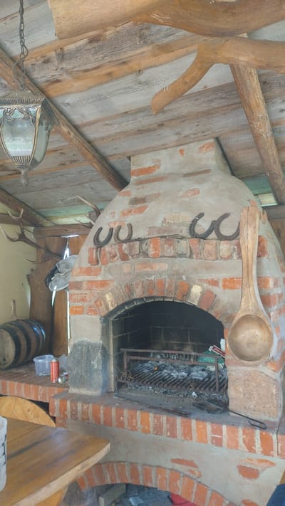 The image depicts a rustic outdoor barbecue area, likely in Kalnrozes, Latvia, featuring a large brick oven with a concrete hood. Several horseshoes are mounted on the hood above the oven's opening. To the left of the oven, a barrel and a clear plastic container with a red aerosol can are visible on a brick ledge. A large wooden ladle hangs from the side of the oven. The ceiling is constructed of wooden beams and planks. In the background, deer antlers are mounted on the wall. The ground appears to be dirt or gravel. The overall scene suggests a functional outdoor cooking space with decorative elements.