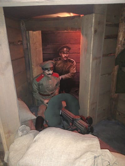 The image displays a museum exhibit simulating a military dugout or trench, located in Мірскі сельскі Савет, Belarus. Two life-sized mannequins, depicted as soldiers, occupy the wooden-planked interior. The mannequin in the left foreground is seated, wearing an olive-drab uniform with red shoulder boards, a matching cap, and a dark mustache. The second mannequin, positioned in the right background, is crouching and also wears an olive-drab uniform and cap. A dark, metallic machine gun, identified by its ribbed barrel shroud and tripod mount, rests on light-colored sandbags in the lower-center of the frame, pointed towards the right. Additional sandbags are stacked along the lower left. The confined space features rough-hewn wooden planks forming the walls and ceiling. The scene is illuminated by dim, red-orange light emanating from the background.