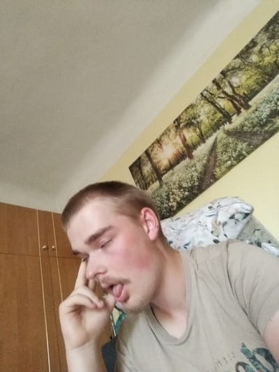 The media file shows a close-up, medium shot of a young Caucasian male in a room, likely in Cēsis, Latvia. The man has short, light brown hair and is wearing a light brown t-shirt. He is looking downwards and to his left, with his eyes partially closed. His right hand is raised to his face, with his index finger touching his temple, and his mouth is slightly open with his tongue visible. His cheeks appear flushed.
The background of the image reveals a wall with a horizontal landscape mural depicting a sunlit forest path lined with white flowers. Above and to the left, a white ceiling is visible. To the left of the man, a section of a wooden wardrobe with two doors and gold-colored knobs is present. A patterned duvet or blanket is visible behind the man's shoulder.
The lighting suggests it is daytime, possibly late afternoon, given the warm glow in the mural. The overall mood seems relaxed or perhaps tired, indicated by the man's pose and facial expression. No visible text is present on the man's clothing or in the immediate surroundings.