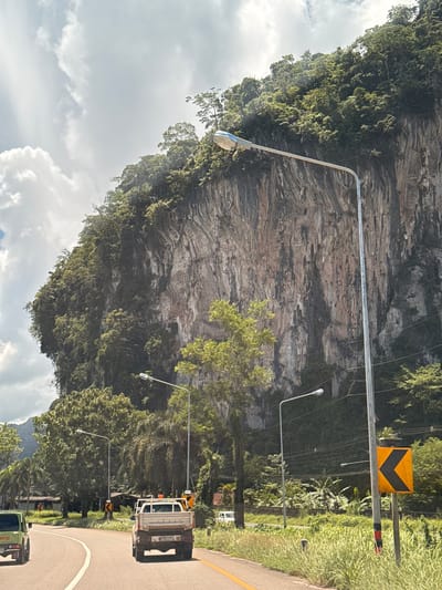 The image shows a rural road winding alongside a sheer, vegetated cliff face. The cliff is composed of layered rock formations, with dense green foliage clinging to its surface. Several streetlights line the road, casting long shadows. A yellow and black chevron road sign indicating a curve is visible. On the right side of the road, a white truck with flashing orange lights is parked. Further down the road, another vehicle, possibly a green utility truck, is partially visible. The sky is filled with dramatic, cloudy formations, suggesting either early morning or late afternoon light. Lush green vegetation borders the road, with tall grass and trees creating a verdant backdrop. The overall scene conveys a sense of natural beauty and rural tranquility, with a hint of roadside maintenance activity. The image appears to be taken from a vehicle moving along the road, given the perspective and the presence of the road itself. There is some text visible on the back of the white truck, but it is illegible. The location is identified as Phang-nga, Thailand.