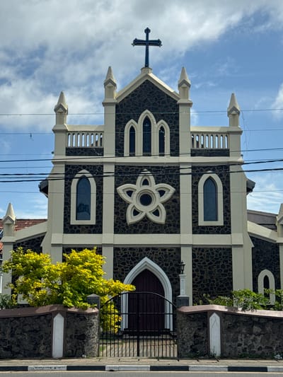 The image shows the front facade of a church in Matara, Sri Lanka. The church has a dark, rough-textured stone exterior with cream-colored trim. It features a central pointed archway leading to a dark wooden door, flanked by arched windows. Above the entrance, a prominent decorative element resembling a five-petaled flower is visible. The roofline is adorned with small spires, and a cross crowns the apex of the central gable. The sky above is partly cloudy. In the foreground, a low stone wall topped with a brown coping encloses a vibrant green tree with yellow foliage, and a black metal gate. A paved sidewalk and road are visible at the bottom of the frame. Electrical wires are strung across the upper portion of the image.