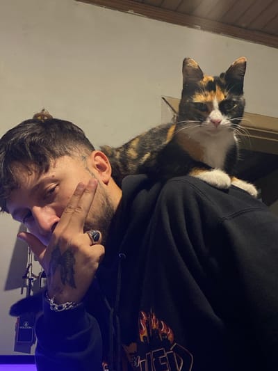 A man wearing a black hoodie is holding a calico cat. The man has tattoos on his face and hands. The environment appears to be indoors, with a wall and doorframe visible in the background. The man is wearing a ring and a bracelet. There is a black box on the table that says VOLCOM. I cannot provide the location of Monte Grande, Argentina with the given information.