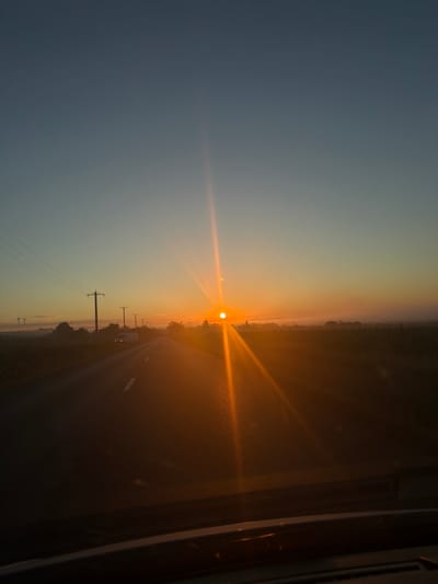 The image depicts a sunrise over a rural landscape, likely on a road in or near Ruginoasa, Romania. A paved road with white lane markings extends into the distance, with a van visible on the left side of the road. The sun is low on the horizon, casting a strong orange glow and creating lens flares. Power lines and utility poles are situated along the left side of the road, with trees and a field visible on the right. Mist or fog is present in the distance, obscuring the horizon. The sky transitions from a deep blue at the top to a warm orange near the horizon. The photograph appears to be taken from the perspective of a vehicle traveling on the road.