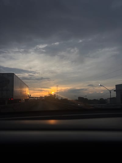 The image depicts a view from inside a vehicle, likely a car, traveling on a road in Tultitlán, Mexico. The sky is dominated by dark, heavy clouds to the left, transitioning to a lighter, hazy horizon towards the center where the sun is setting or rising. A large semi-trailer truck is visible on the road, partially obstructing the view. Power lines stretch across the upper portion of the frame, intersecting with streetlights along the roadside. The general environment suggests an industrial or highway setting, with elements of urban infrastructure. The image captures a moment of transition in lighting conditions, perhaps at dawn or dusk.