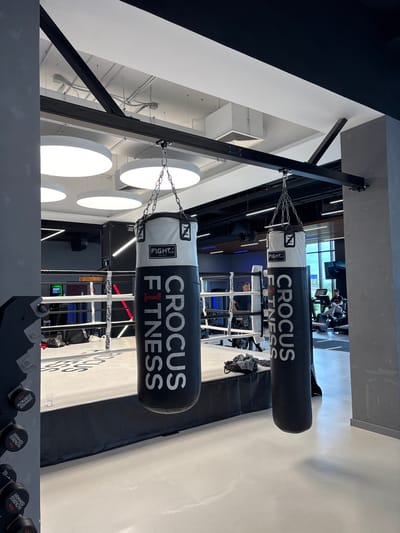 The image depicts the interior of a boxing gym, likely in the city of Monolit, Russia. In the foreground, two large punching bags hang from a metal frame. The bag on the right is predominantly black with white lettering that reads "CROCUS FITNESS." The bag on the left is white with black lettering. A boxing ring is visible in the middle ground, with white ropes and a raised platform. Inside the ring, a person wearing dark clothing is seen standing. In the background, exercise equipment, including treadmills, is present, and several individuals can be observed using the machines. The gym features modern lighting, including circular ceiling lights, and industrial-style exposed beams and ductwork. The overall environment suggests a well-equipped fitness facility.