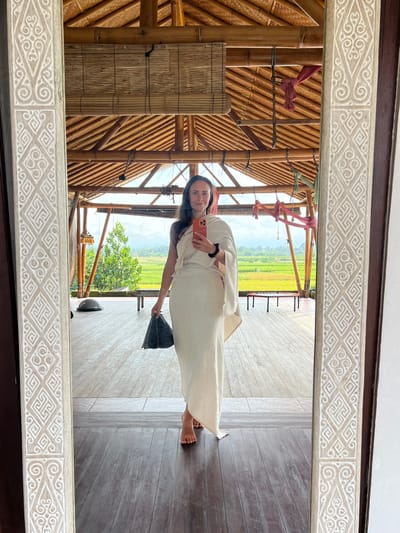 A woman is taking a mirror selfie. She is wearing a light-colored dress. She is holding a mobile phone in her right hand and a small bag in her left hand. The mirror reflects a bamboo structure with a thatched roof. In the background, there is a view of a rice field. The location is Singakerta, Indonesia.