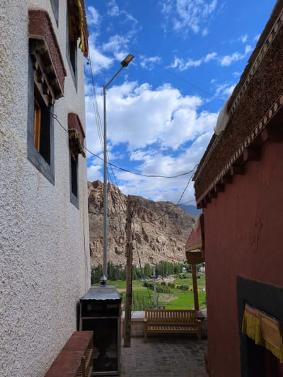 The image shows a narrow alleyway between two buildings, with a mountainous landscape in the background. The building on the left is white with gray and maroon trim around windows. The building on the right is red with a decorative, textured roofline. A wooden bench sits in the alley, and beyond it, a green field is visible with trees and a village nestled at the base of the mountains. A lamppost stands near the white building. The sky is blue with scattered white clouds. The scene appears to be located in a high-altitude, possibly arid region.