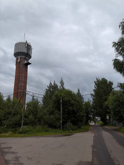 The image depicts a wide, asphalt road that curves to the right, disappearing into a cluster of trees. The road is wet in patches, suggesting recent rain. On the left side of the road, dense green foliage forms a wall, with a tall, red brick water tower rising above it in the distance. The water tower has a gray, cylindrical upper section and appears to have antennas and ladders attached to its exterior. On the right side of the road, more trees and vegetation are visible, along with a few utility poles and overhead wires. A single person is walking away from the camera down the road. The sky is overcast with heavy, gray clouds. The scene is set in Maloye Verevo, Russia.