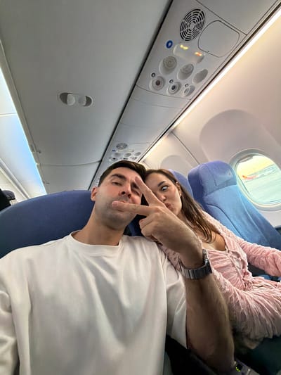 A man and a woman are on an airplane. The man is wearing a white shirt and a watch on his left wrist and is making a peace sign with his right hand near his face. The woman is wearing a pink top. An airplane window is visible in the upper left portion of the image. It is daytime, and an airport structure is visible outside of the window. The location is an airplane cabin, possibly landing at or departing from Beijing Capital International Airport in the Shunyi District, China.