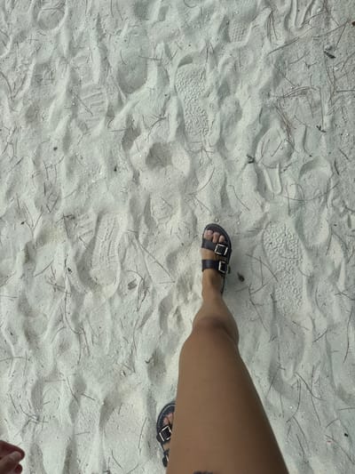 The image depicts an overhead view of an individual's legs and feet on a light-colored sandy beach in Berry Islands, Bahamas. The subject wears dark brown sandals, each secured with two metallic buckles. The right leg, extending into the mid-right of the frame, shows the sandal in profile, while a portion of the left foot with its sandal is visible at the bottom-left. The fine-grained sand is characterized by numerous undulations and multiple distinct footprints, one clearly displaying a ridged sole pattern. Scattered throughout the sand are thin, dark, dry organic debris, resembling pine needles. A hand is partially visible in the lower-left corner of the frame.